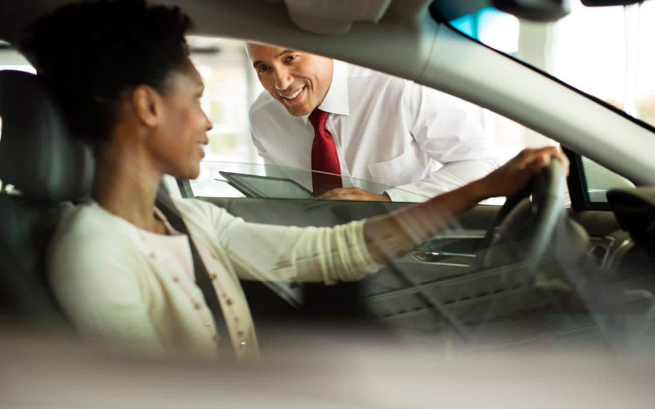 Car salesperson assisting a woman inside a vehicle at a dealership.