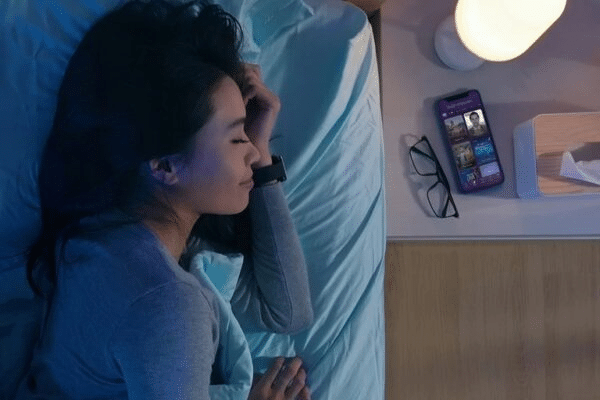 Woman sleeping peacefully beside smartphone with open streaming app; cozy nighttime scene.
