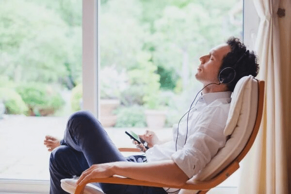 Man relaxing in a chair by a window, listening to music with headphones, holding a smartphone.