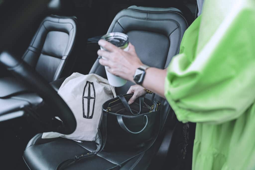 A person in a green outfit reaching into a black handbag inside a car, holding a coffee cup in the other hand, with a canvas bag featuring a logo resting on the seat.
