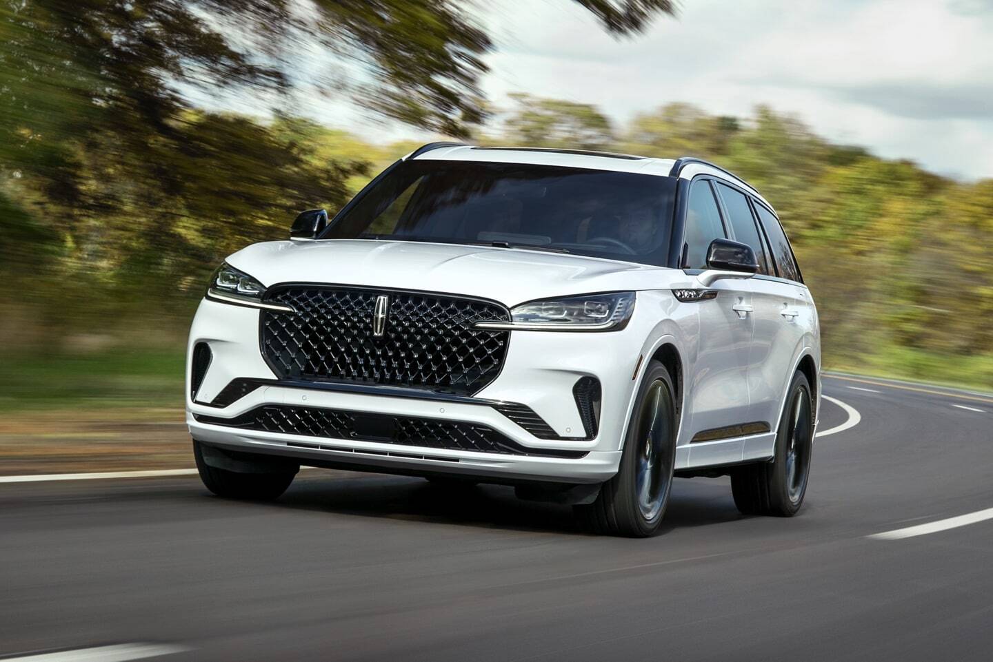 A white luxury SUV with a distinctive front grille and modern design, captured in motion on a curved road surrounded by autumn foliage.