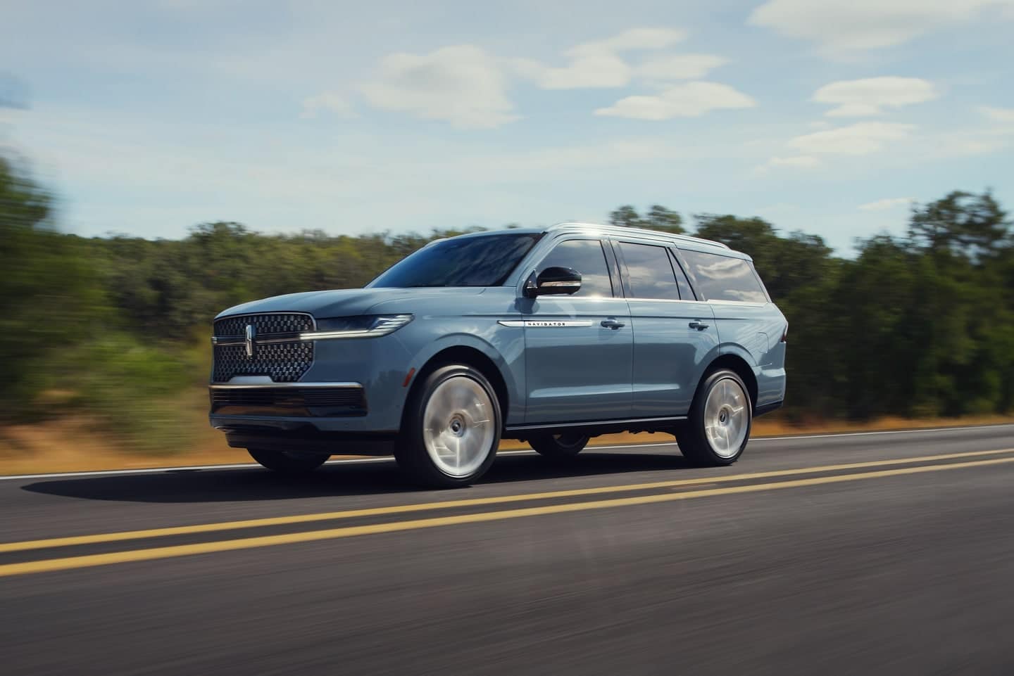 A sleek blue luxury SUV driving on a countryside road, highlighting its modern design and performance features against a backdrop of greenery and blue sky.